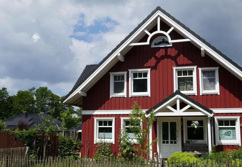 Realistische Illustration eines idyllischen Sommerhauses inmitten üppiger Natur, mit einer Familie bei typischen Sommeraktivitäten wie Grillen und Spielen im Garten, Hauptfarbton Rot.