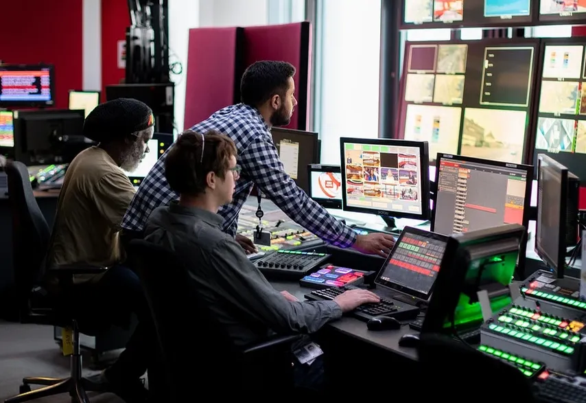 Heike Greis als deutsche Fernsehmoderatorin in einem professionellen Studio mit roten Farbtönen, umgeben von Kameras und Monitoren, hält ein Mikrofon in der Hand.